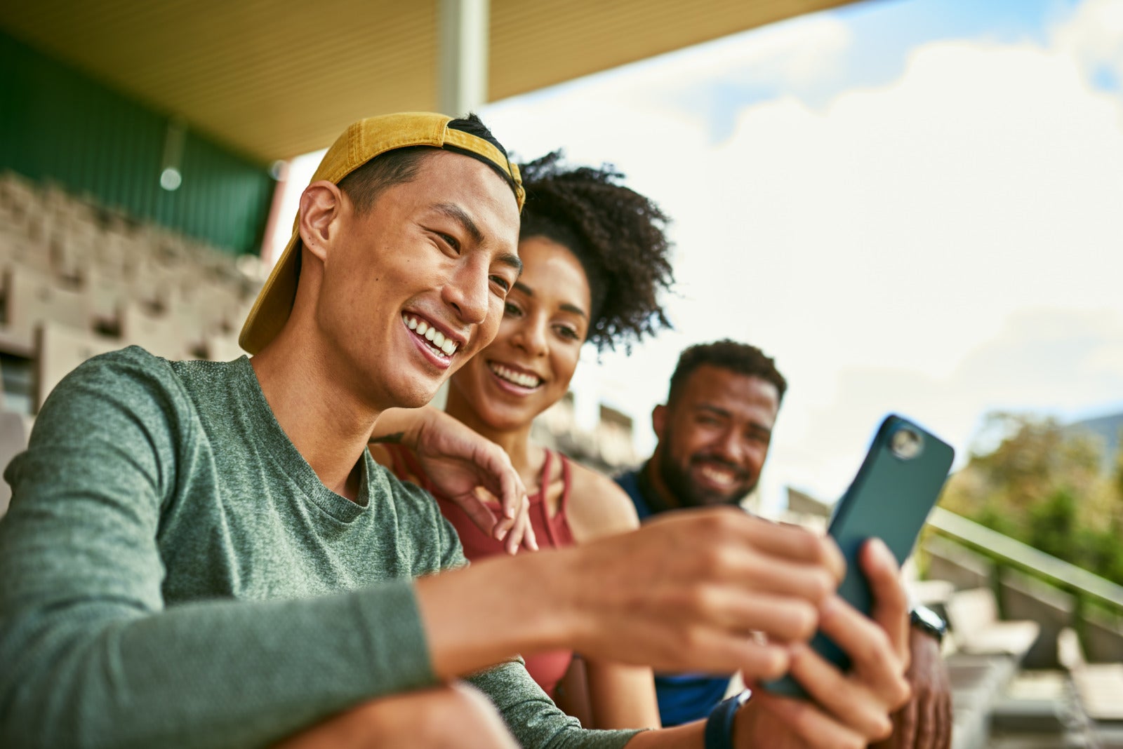 People outdoors smiling at phone, dressed casually. Young, diverse group looking and laughing at website on mobile excitedly. Friends sitting with each other on stadium chairs