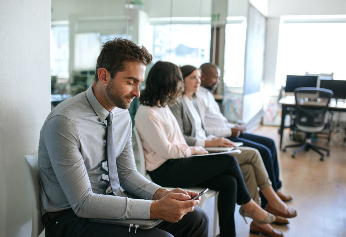 Young man smiling at his phone while waiting with other interviewees