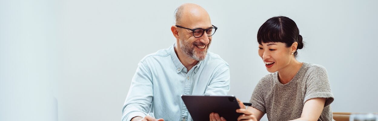 Two colleagues are sitting in a modern office while looking at information on a tablet and smiling.