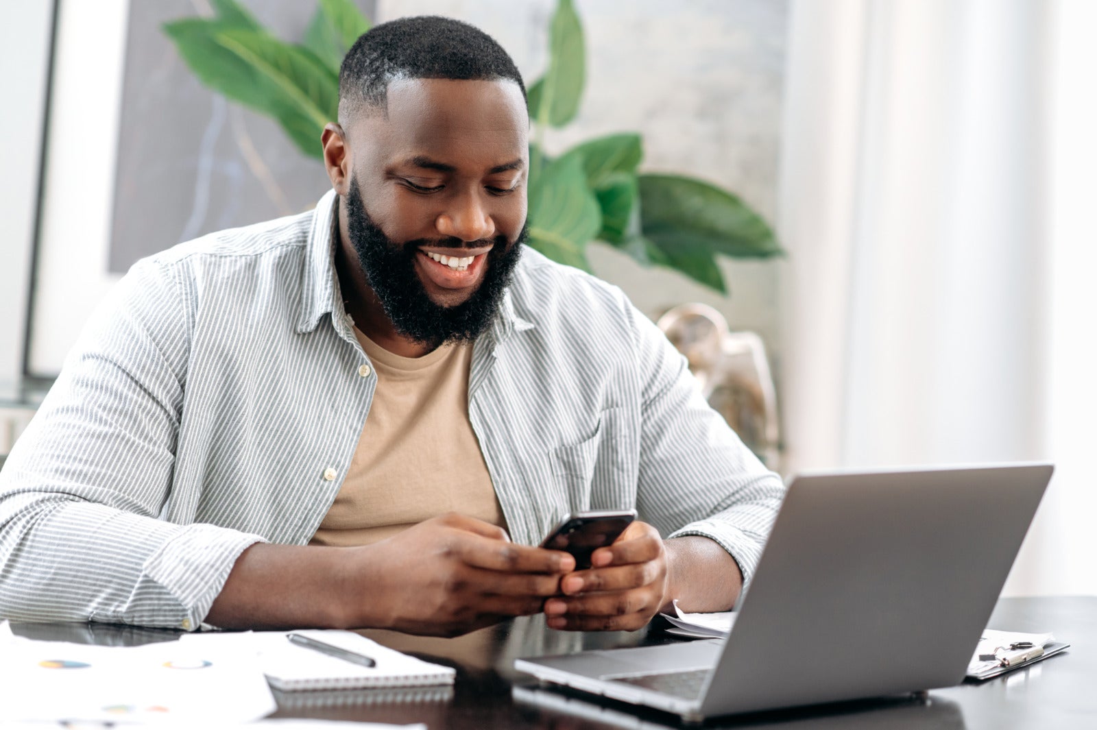 man sitting at a desk in the office, using his smartphone, in front of laptop, smiling