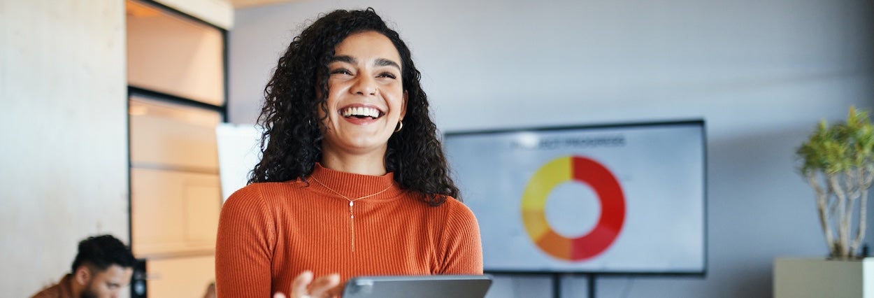 Smiling businesswoman with curly hair presents data on a tablet. A pie chart is displayed on a screen behind her. A colleague is working in the background, creating a collaborative office environment.