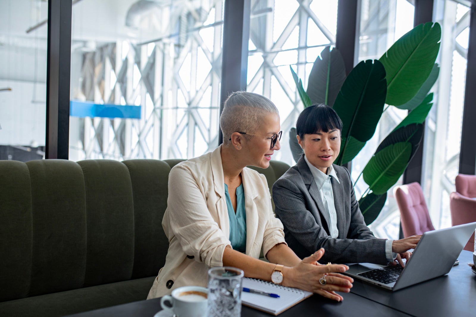 Two businesswomen in professional attire smile and discuss work while reviewing information on a laptop in a modern office setting, emphasizing cooperation and productivity