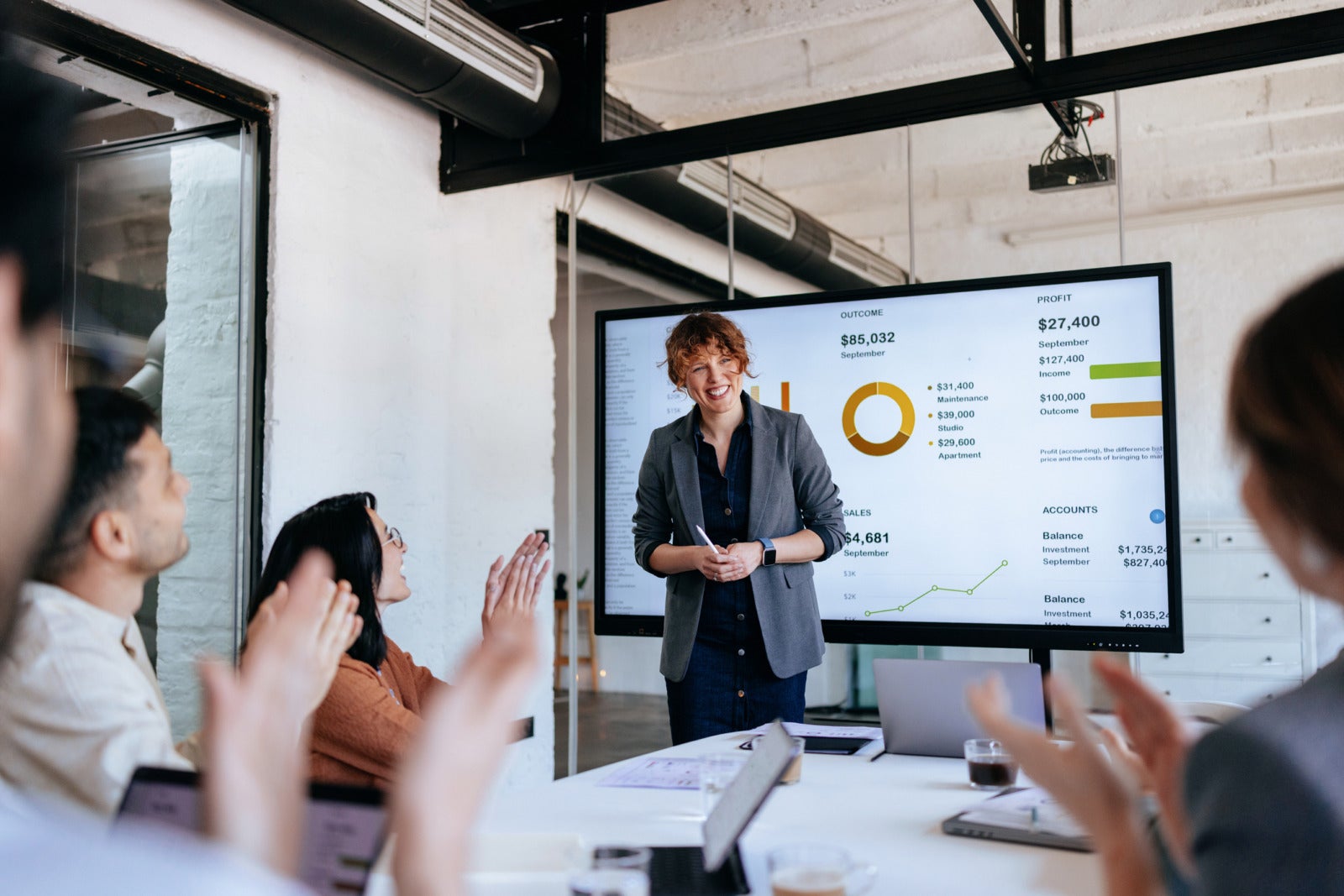 A professional presenter stands beside a large screen displaying charts and graphs while colleagues applaud and engage in a dynamic business meeting in a bright, modern office space.